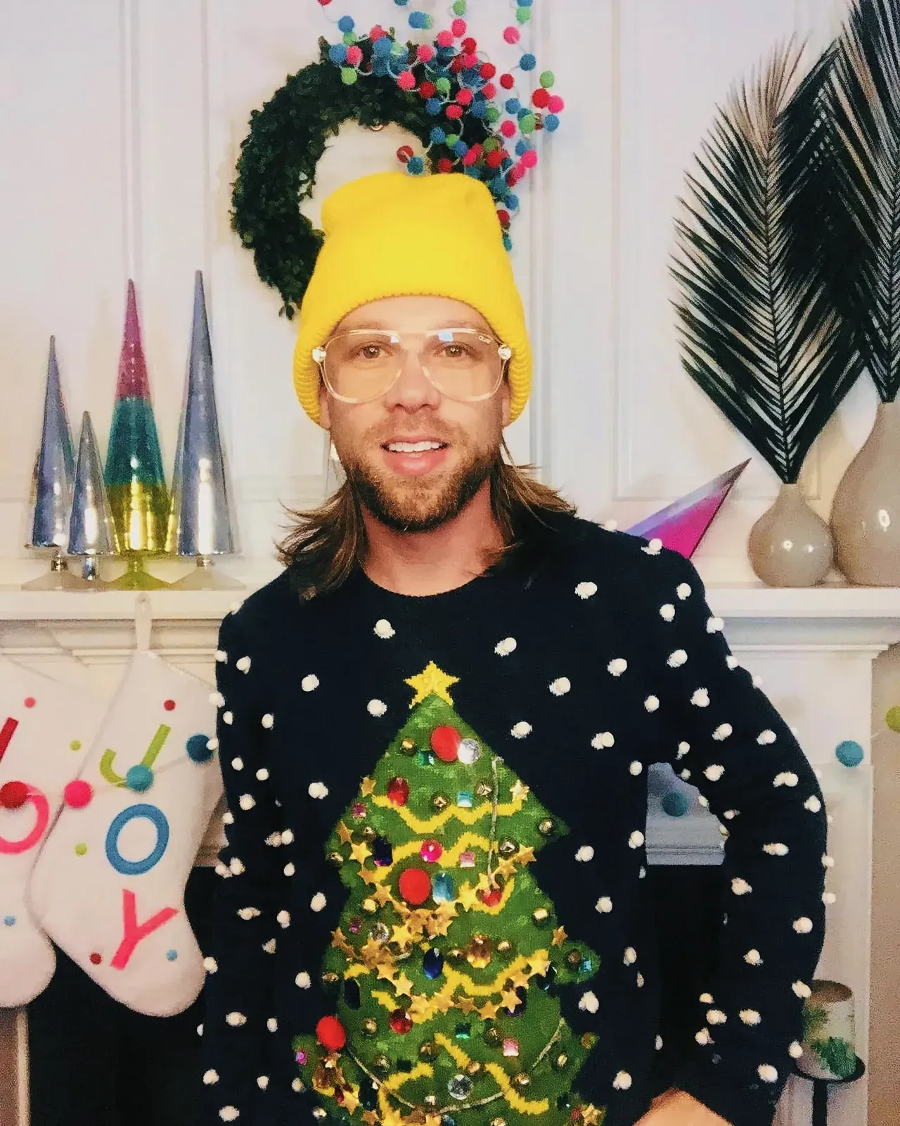 The Ugly Christmas Sweater Phenomenon: A man wearing a vibrant yellow hat and glasses, smiling in front of a Christmas tree with a DIY sweater.
