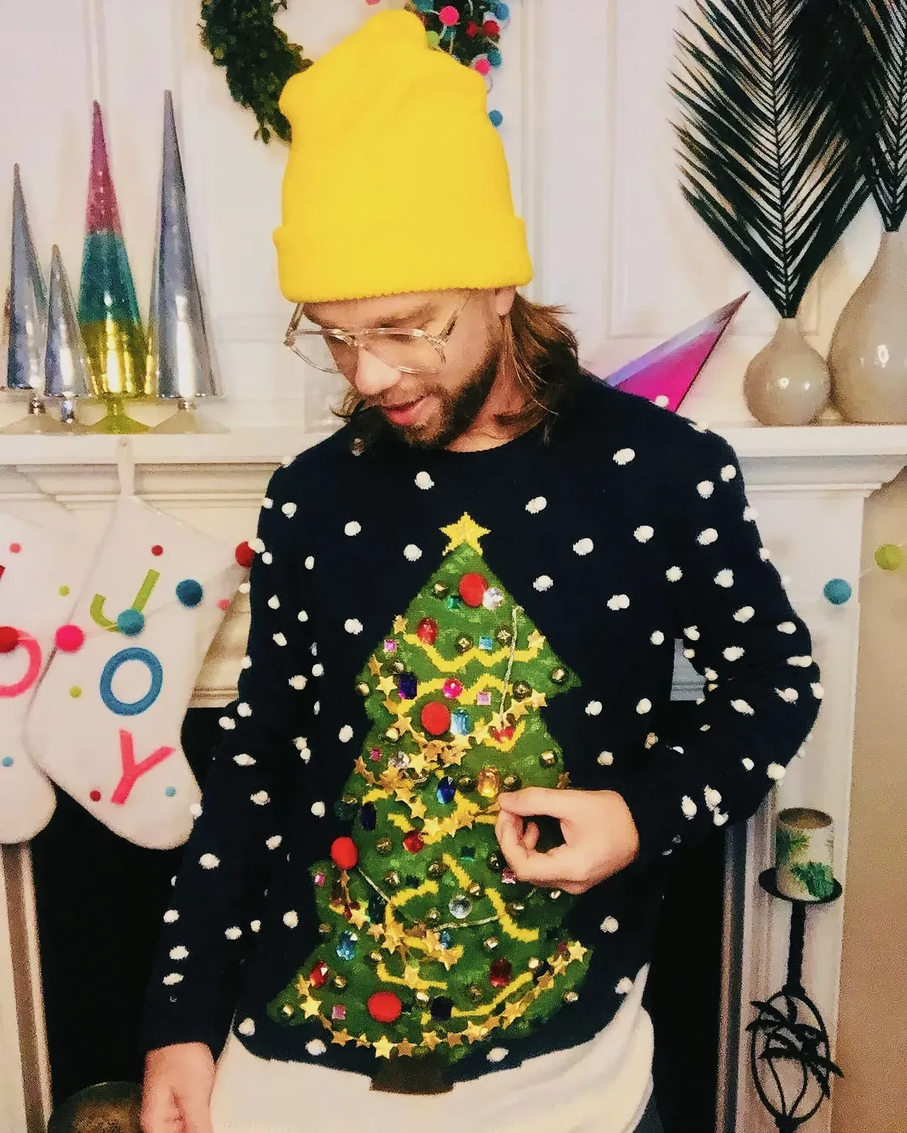 Man wearing a DIY ugly Christmas sweater and yellow hat with pom-poms in a festive indoor setting with a Christmas tree and decorations.